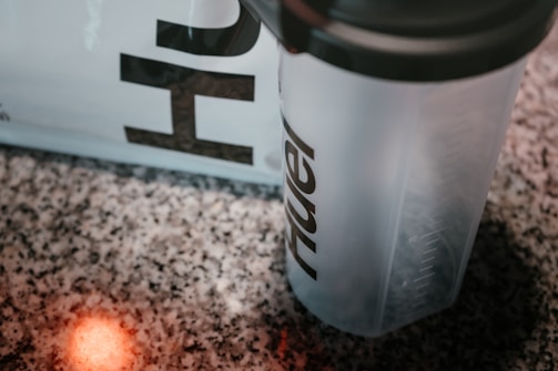 A close-up view of a shaker bottle with the visible partial text on it, placed on a speckled countertop. A soft, red light reflection is visible on the counter.