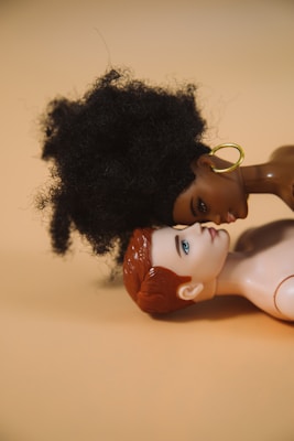 Two dolls are positioned lying down, facing each other closely. One doll has curly black hair and is adorned with large gold hoop earrings, while the other doll has short, red hair and fair skin. They are set against a neutral, beige background.