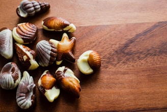 A collection of assorted seashell-shaped chocolates rests on a wooden surface. The chocolates feature marbled patterns in various shades of brown and white, resembling seashell designs.