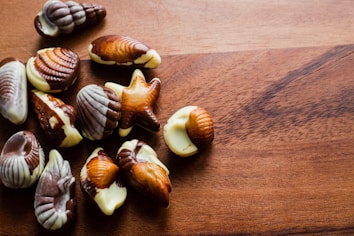 A collection of assorted seashell-shaped chocolates rests on a wooden surface. The chocolates feature marbled patterns in various shades of brown and white, resembling seashell designs.