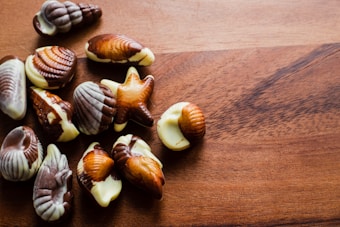 A collection of assorted seashell-shaped chocolates rests on a wooden surface. The chocolates feature marbled patterns in various shades of brown and white, resembling seashell designs.