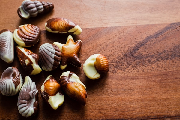 A collection of assorted seashell-shaped chocolates rests on a wooden surface. The chocolates feature marbled patterns in various shades of brown and white, resembling seashell designs.