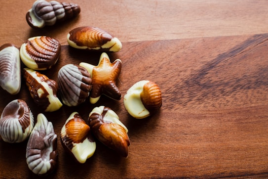 A collection of assorted seashell-shaped chocolates rests on a wooden surface. The chocolates feature marbled patterns in various shades of brown and white, resembling seashell designs.