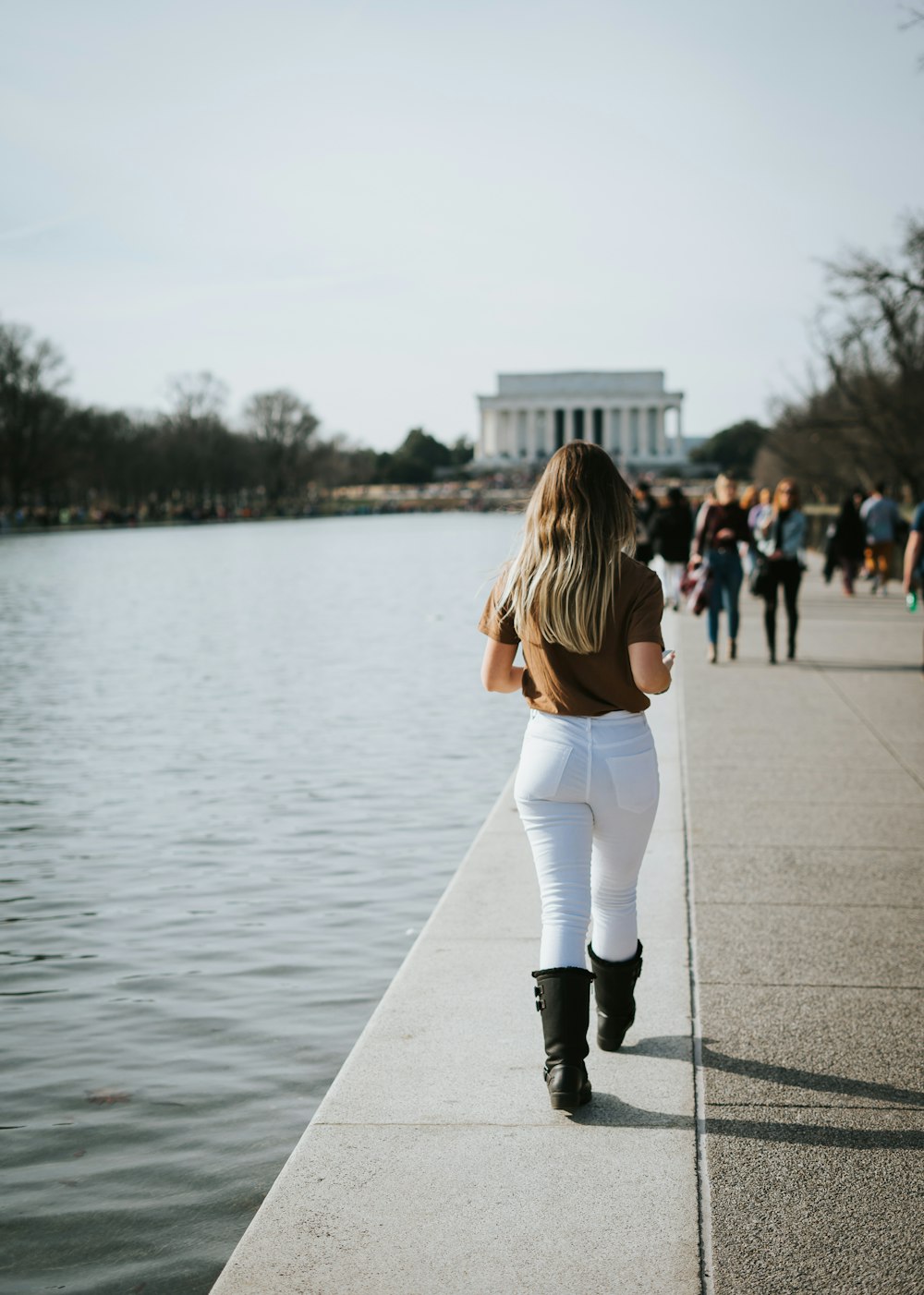 woman in white long sleeve shirt and white pants walking on sidewalk near body of water