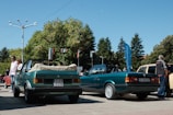 Two green convertible cars are parked in an outdoor area with trees and flags in the background. There are several people standing around, including one man who appears to be inspecting one of the cars. The day is sunny with a clear blue sky.