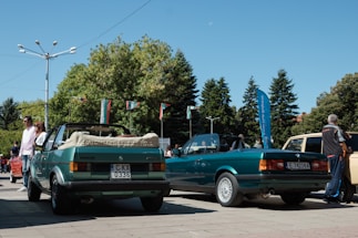 Two green convertible cars are parked in an outdoor area with trees and flags in the background. There are several people standing around, including one man who appears to be inspecting one of the cars. The day is sunny with a clear blue sky.