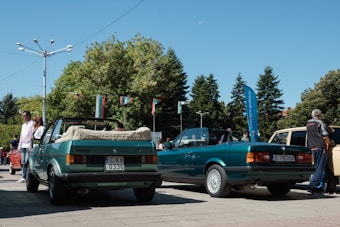 Two green convertible cars are parked in an outdoor area with trees and flags in the background. There are several people standing around, including one man who appears to be inspecting one of the cars. The day is sunny with a clear blue sky.