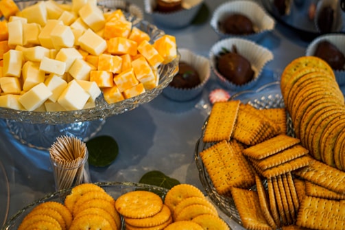 A display of assorted cheeses and crackers on a table. Cubes of white and orange cheese are arranged on a crystal platter, surrounded by stacks of round and square crackers. Toothpicks are placed nearby for serving. Chocolate-covered items in paper cups are visible in the background.