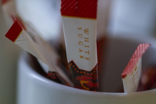 Several white and red sugar packets are placed upright in a white container. The packets have the words 'white sugar' printed on them and feature a decorative design near the bottom. The focus is on the packets, with the background softly blurred.