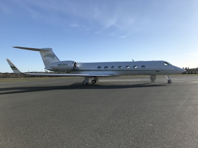 A sleek, modern private jet with a streamlined design is stationary on a tarmac under a clear blue sky. The aircraft is white with green accents and features several circular windows along its body.