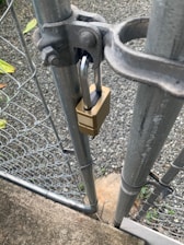 A metal gate is secured with a brass padlock. The gate is part of a chain-link fence, and is attached to a sturdy metal post. A gravel path and some greenery are visible in the background.