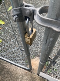 A metal gate is secured with a brass padlock. The gate is part of a chain-link fence, and is attached to a sturdy metal post. A gravel path and some greenery are visible in the background.