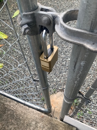 A metal gate is secured with a brass padlock. The gate is part of a chain-link fence, and is attached to a sturdy metal post. A gravel path and some greenery are visible in the background.