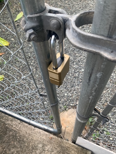 A metal gate is secured with a brass padlock. The gate is part of a chain-link fence, and is attached to a sturdy metal post. A gravel path and some greenery are visible in the background.
