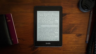 A Kindle displaying text is placed on a brown wooden table. To the left, there is a closed black book or journal and a smaller red book, while on the right, a lens cap and a pen are visible.