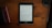 A Kindle displaying text is placed on a brown wooden table. To the left, there is a closed black book or journal and a smaller red book, while on the right, a lens cap and a pen are visible.