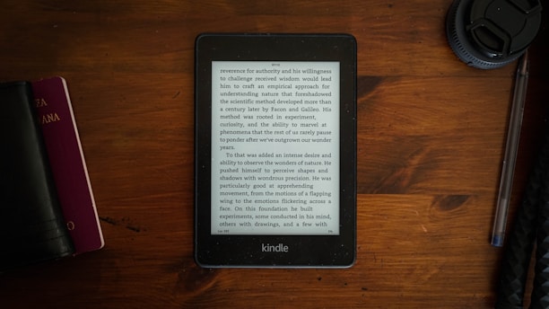 A Kindle displaying text is placed on a brown wooden table. To the left, there is a closed black book or journal and a smaller red book, while on the right, a lens cap and a pen are visible.