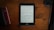 A Kindle displaying text is placed on a brown wooden table. To the left, there is a closed black book or journal and a smaller red book, while on the right, a lens cap and a pen are visible.