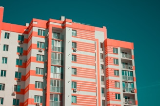 A modern multi-story apartment building features a distinctive pattern of coral and white stripes across its facade. The building has numerous windows and balconies, with air conditioning units mounted on the outer walls. The sky is clear, providing a contrasting teal background.