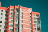 A modern multi-story apartment building features a distinctive pattern of coral and white stripes across its facade. The building has numerous windows and balconies, with air conditioning units mounted on the outer walls. The sky is clear, providing a contrasting teal background.
