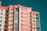 A modern multi-story apartment building features a distinctive pattern of coral and white stripes across its facade. The building has numerous windows and balconies, with air conditioning units mounted on the outer walls. The sky is clear, providing a contrasting teal background.