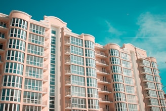 A modern multi-story residential building with curved balconies and large glass windows against a clear blue sky. The architecture features soft peach-colored walls and symmetrical design elements.
