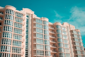 A modern multi-story residential building with curved balconies and large glass windows against a clear blue sky. The architecture features soft peach-colored walls and symmetrical design elements.