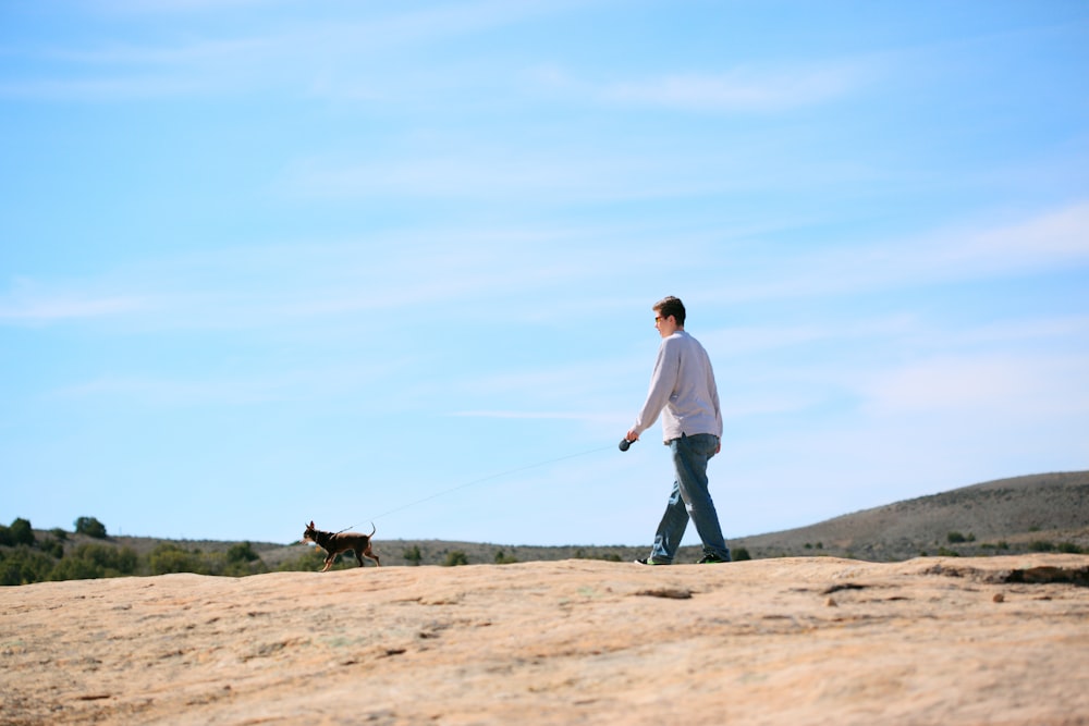 白い長袖シャツと黒いズボンを着た男が茶色の野原で黒い犬と一緒に歩いている