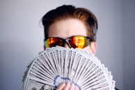 A person wearing reflective sunglasses is holding a large fan of hundred-dollar bills. The background is plain and unadorned, and the focus is on the money and the sunglasses.