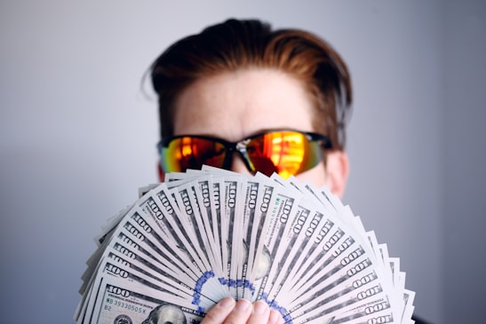 A person wearing reflective sunglasses is holding a large fan of hundred-dollar bills. The background is plain and unadorned, and the focus is on the money and the sunglasses.