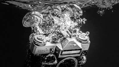 A vintage Zenit-E camera is submerged in water, creating a dramatic splash with bubbles surrounding it. The image is in black and white, highlighting the textures and details of the camera and the water.