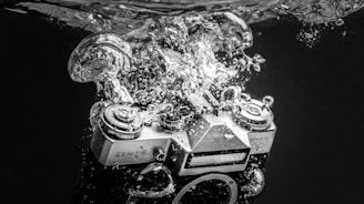 A vintage Zenit-E camera is submerged in water, creating a dramatic splash with bubbles surrounding it. The image is in black and white, highlighting the textures and details of the camera and the water.