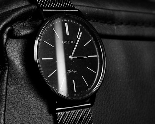 A classic analog wristwatch with a black watch face and minimalist design. The watch face has sleek white hour markers and slender hands, complemented by a mesh strap. The watch rests on a textured leather surface.