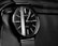 A classic analog wristwatch with a black watch face and minimalist design. The watch face has sleek white hour markers and slender hands, complemented by a mesh strap. The watch rests on a textured leather surface.