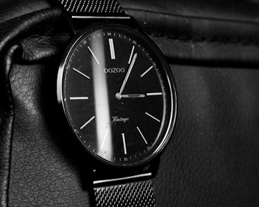 A classic analog wristwatch with a black watch face and minimalist design. The watch face has sleek white hour markers and slender hands, complemented by a mesh strap. The watch rests on a textured leather surface.