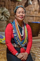 A person wears traditional attire adorned with colorful beaded necklaces and large metal discs. The background features woven walls and a basket, suggesting a cultural or indigenous setting. The person is seated on a wooden floor.