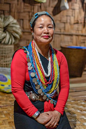A person wears traditional attire adorned with colorful beaded necklaces and large metal discs. The background features woven walls and a basket, suggesting a cultural or indigenous setting. The person is seated on a wooden floor.
