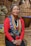 A person wears traditional attire adorned with colorful beaded necklaces and large metal discs. The background features woven walls and a basket, suggesting a cultural or indigenous setting. The person is seated on a wooden floor.