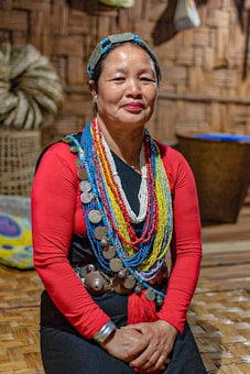 A person wears traditional attire adorned with colorful beaded necklaces and large metal discs. The background features woven walls and a basket, suggesting a cultural or indigenous setting. The person is seated on a wooden floor.