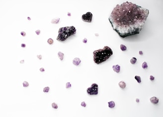 An assortment of amethyst crystals is scattered across a white surface. The arrangement includes various sizes and shapes, including raw chunks and heart-shaped crystals. The colors range from deep purple to light lavender. Some crystals display a sparkling, gem-like quality.