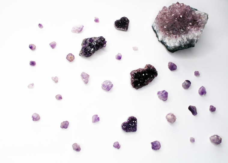 An assortment of amethyst crystals is scattered across a white surface. The arrangement includes various sizes and shapes, including raw chunks and heart-shaped crystals. The colors range from deep purple to light lavender. Some crystals display a sparkling, gem-like quality.