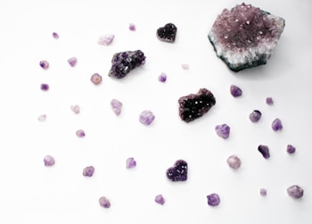 An assortment of amethyst crystals is scattered across a white surface. The arrangement includes various sizes and shapes, including raw chunks and heart-shaped crystals. The colors range from deep purple to light lavender. Some crystals display a sparkling, gem-like quality.