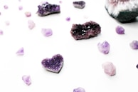 Various amethyst crystals are scattered across a white background, including one shaped like a heart. The crystals have a rich purple color and are in different sizes and shapes, ranging from small shards to larger clusters with a crystalline texture.