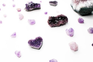 Various amethyst crystals are scattered across a white background, including one shaped like a heart. The crystals have a rich purple color and are in different sizes and shapes, ranging from small shards to larger clusters with a crystalline texture.