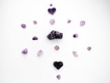 Several purple crystals and stones are arranged in a circular pattern on a white background. The stones vary in shape and size, with two heart-shaped crystal clusters positioned at the top and bottom.