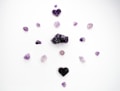 Several purple crystals and stones are arranged in a circular pattern on a white background. The stones vary in shape and size, with two heart-shaped crystal clusters positioned at the top and bottom.