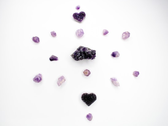 Several purple crystals and stones are arranged in a circular pattern on a white background. The stones vary in shape and size, with two heart-shaped crystal clusters positioned at the top and bottom.