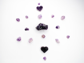 Several purple crystals and stones are arranged in a circular pattern on a white background. The stones vary in shape and size, with two heart-shaped crystal clusters positioned at the top and bottom.