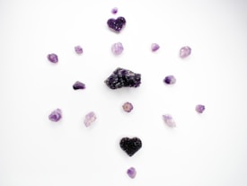Several purple crystals and stones are arranged in a circular pattern on a white background. The stones vary in shape and size, with two heart-shaped crystal clusters positioned at the top and bottom.