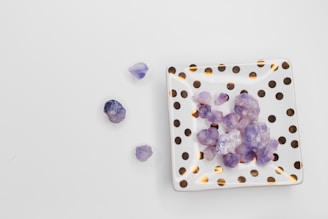 Purple crystals rest on a square, white dish decorated with gold polka dots, placed on a plain white surface. Several crystals are scattered beside the dish, showing irregular shapes and translucent qualities.